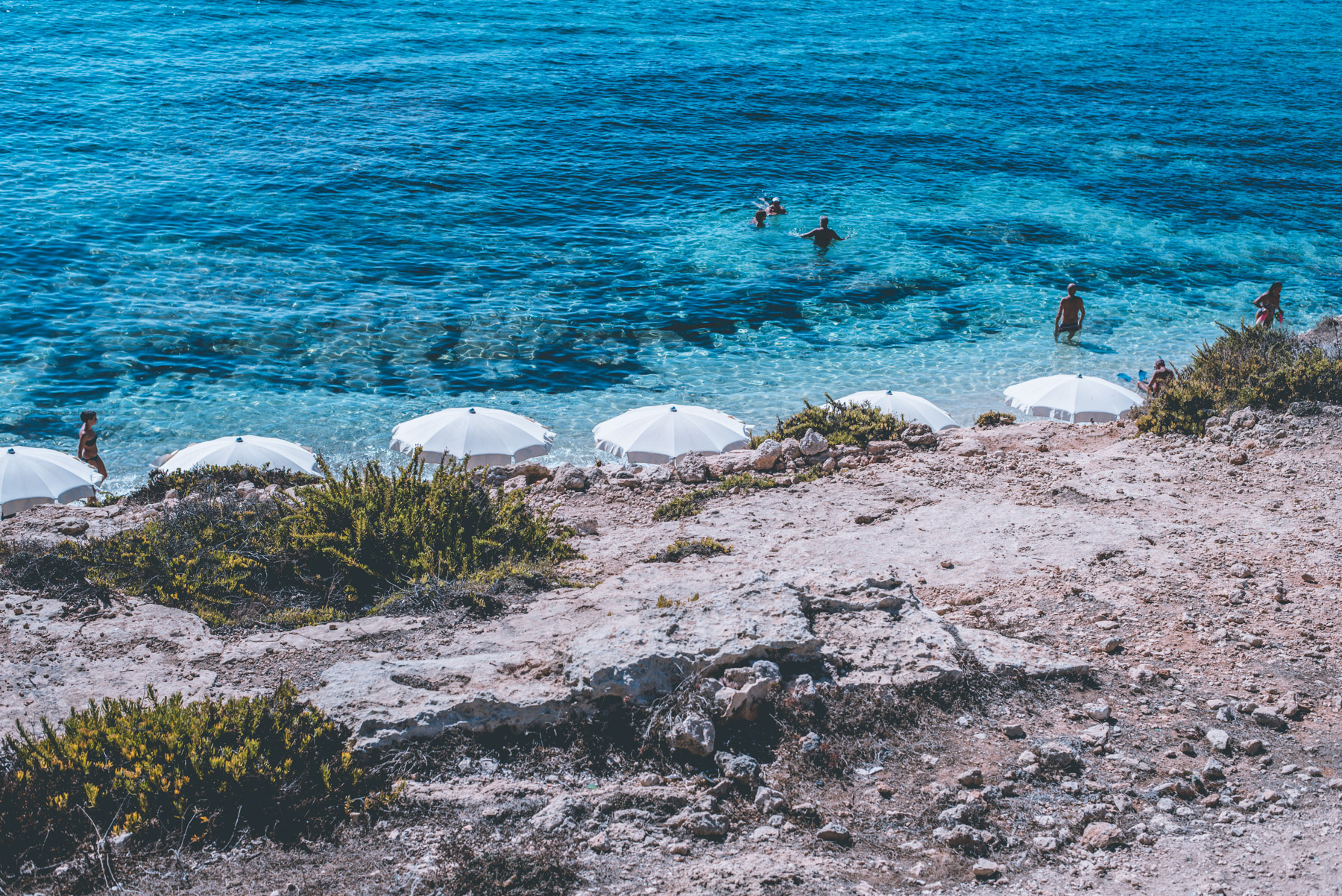 view from above of a small beach on the island of Lampedusa with blue sea and some white beach umbrellas and people swimming in the sea
