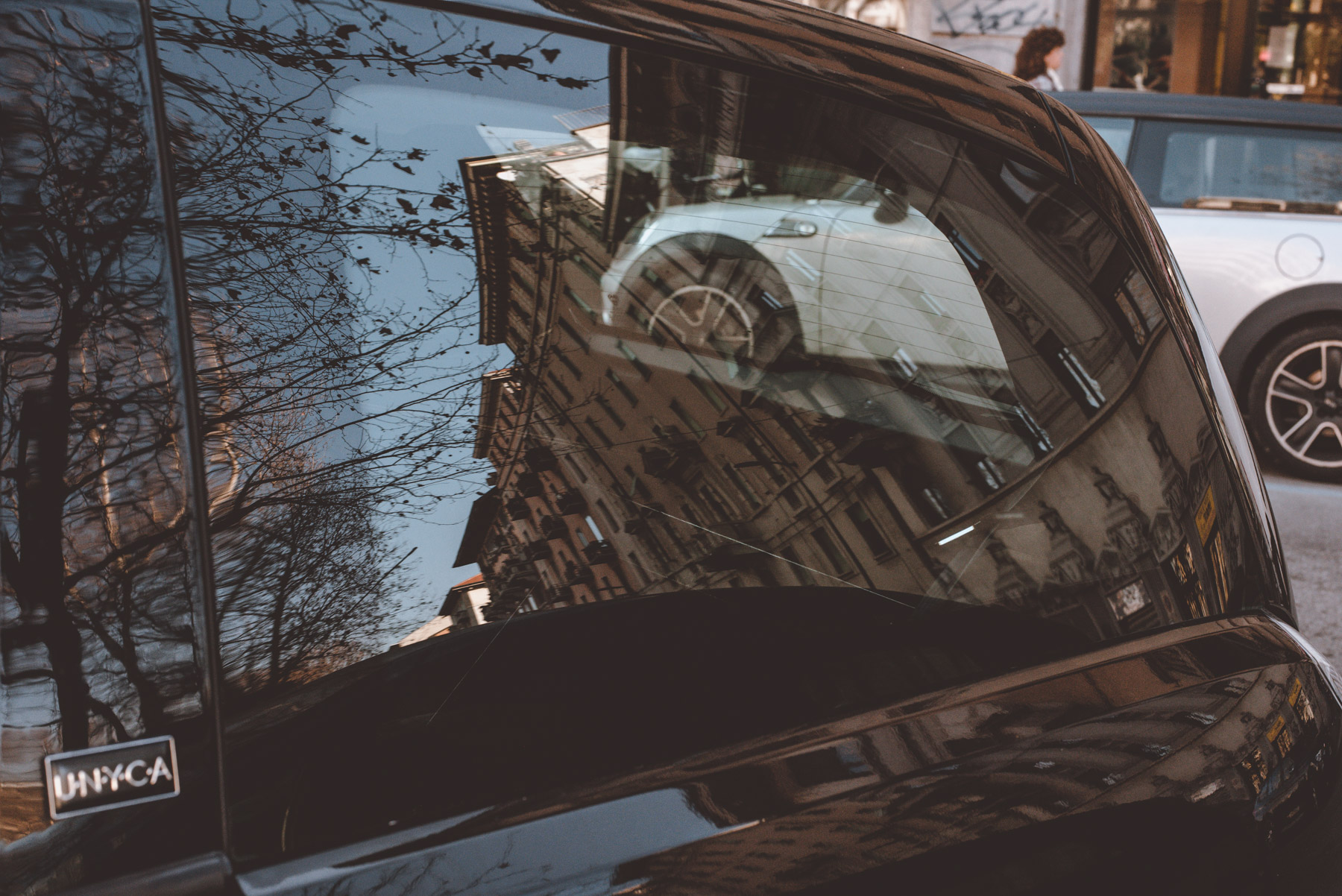 A street photograph showing reflections of a car some buildings and trees on the streets of Milano